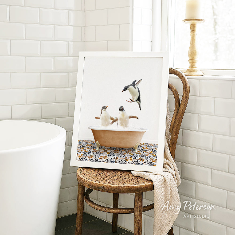 A framed image of two penguins sitting in a bathtub, surrounded by a patterned tile design, is displayed on a wooden chair in a bathroom setting.