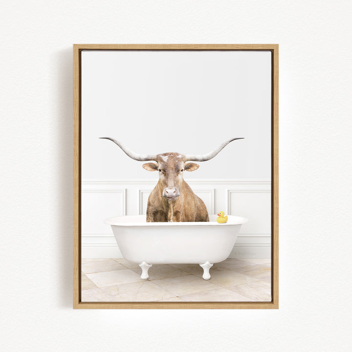 A brown cow with large horns is sitting in a white bathtub, with a yellow rubber duck floating in the water.