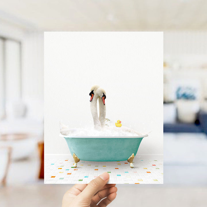 A hand holds up a framed image depicting a white swan standing in a blue bathtub, with a yellow rubber duck floating nearby.