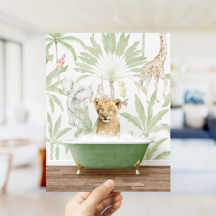 A hand holds up a framed print featuring a baby lion sitting in a green bathtub surrounded by tropical foliage, including a giraffe and an elephant.
