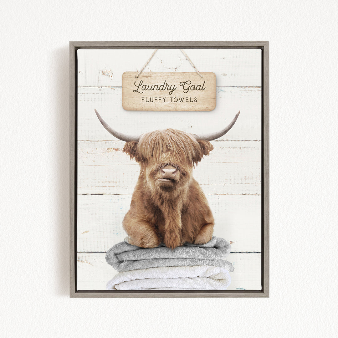 A framed image of a brown cow with long horns sitting on a stack of fluffy towels, with a sign above that says "Laundry Goal Fluffy Towels".