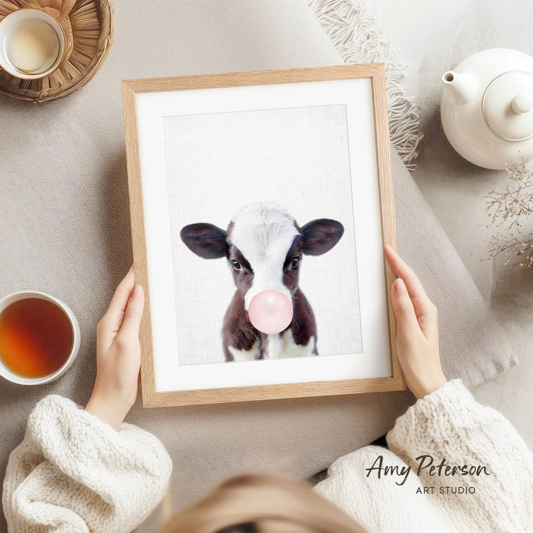 A person is holding a framed picture of a cow blowing a bubble with a pink bubble gum bubble, surrounded by various objects such as a teapot, a cup, and a bowl.