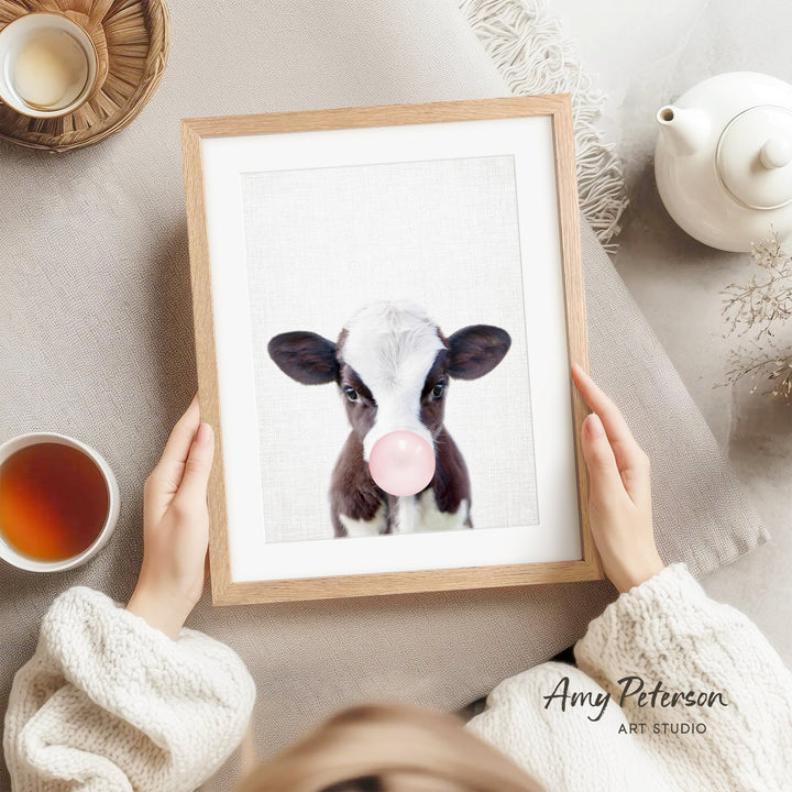 A person is holding a framed picture of a cow blowing a bubble with a pink bubble gum bubble, surrounded by various objects such as a teapot, a cup, and a bowl.