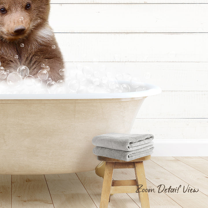 A brown bear is sitting in a bathtub filled with bubbles, with a towel and stool nearby.