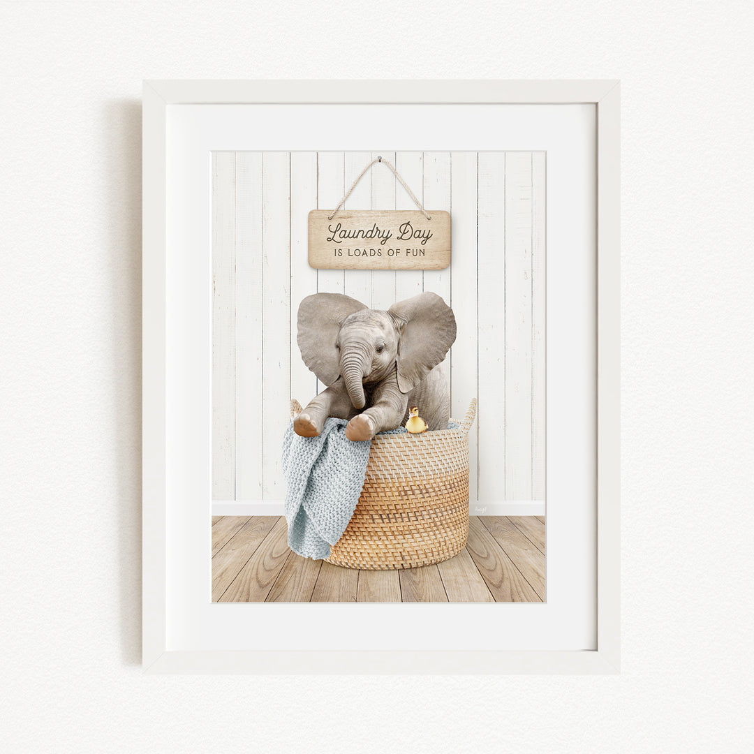 A framed image of a baby elephant sitting in a woven basket, with a sign above it that says "Laundry Day is loads of fun".