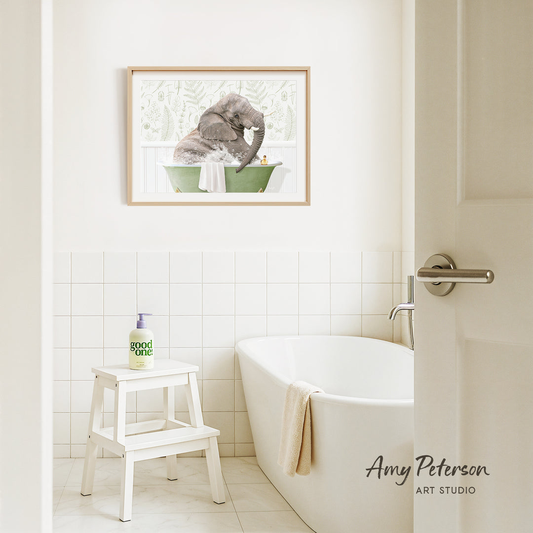 A framed painting of an elephant in a bathtub hangs on a white tiled wall above a white bathtub in a bathroom.