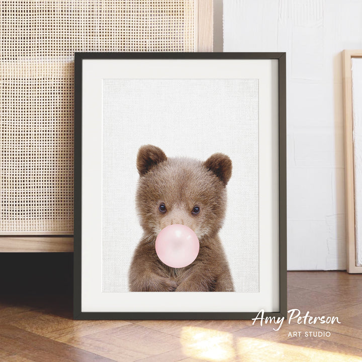 A framed photograph of a brown bear blowing a bubble with a pink bubble gum bubble.