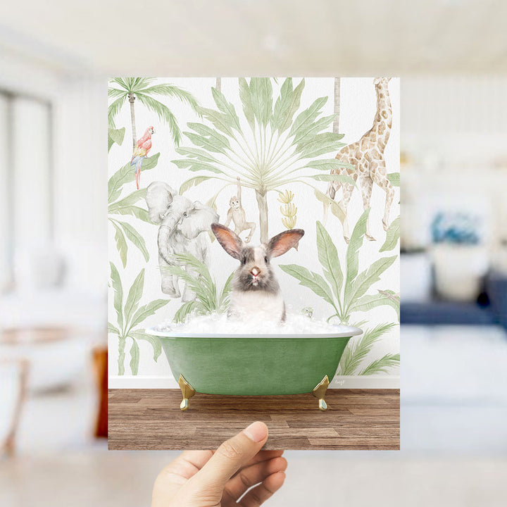 A hand holds up a framed print featuring a whimsical scene of a rabbit sitting in a green bathtub surrounded by tropical foliage, including a giraffe and other animals.