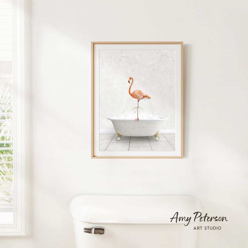 A framed print of a flamingo standing in a white bathtub is hanging on a white wall.