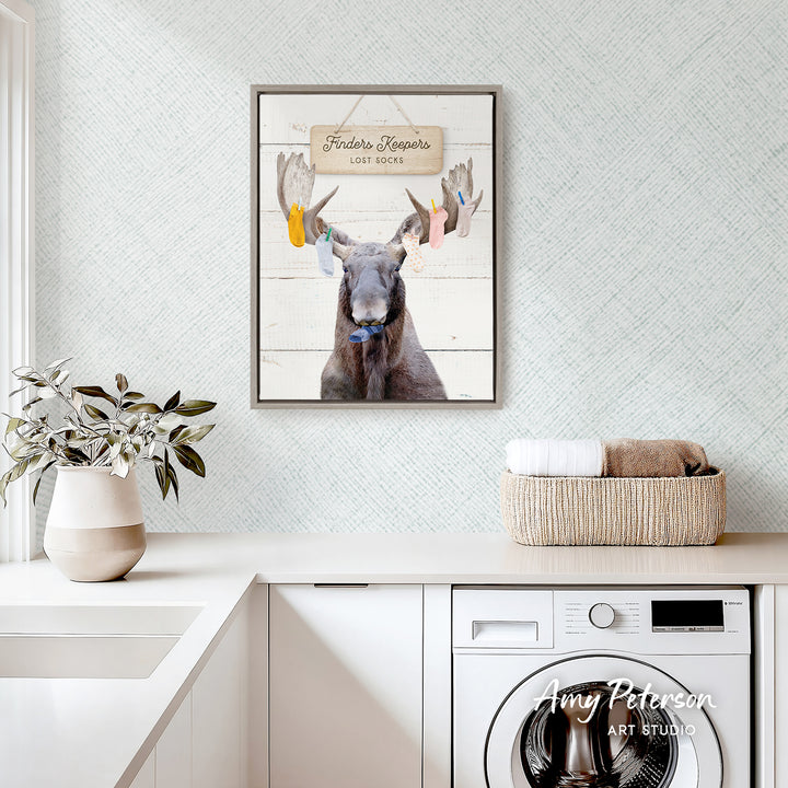 a framed photo of a moose with antlers, hanging on a wall above a white countertop with a basket and a plant.
