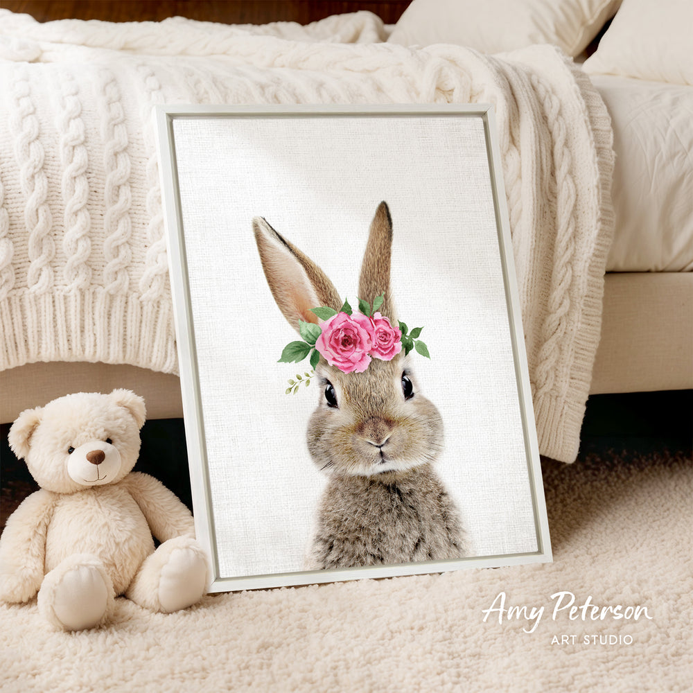 A framed print of a rabbit wearing a pink flower crown is displayed on a white bedspread, accompanied by a teddy bear on the floor.