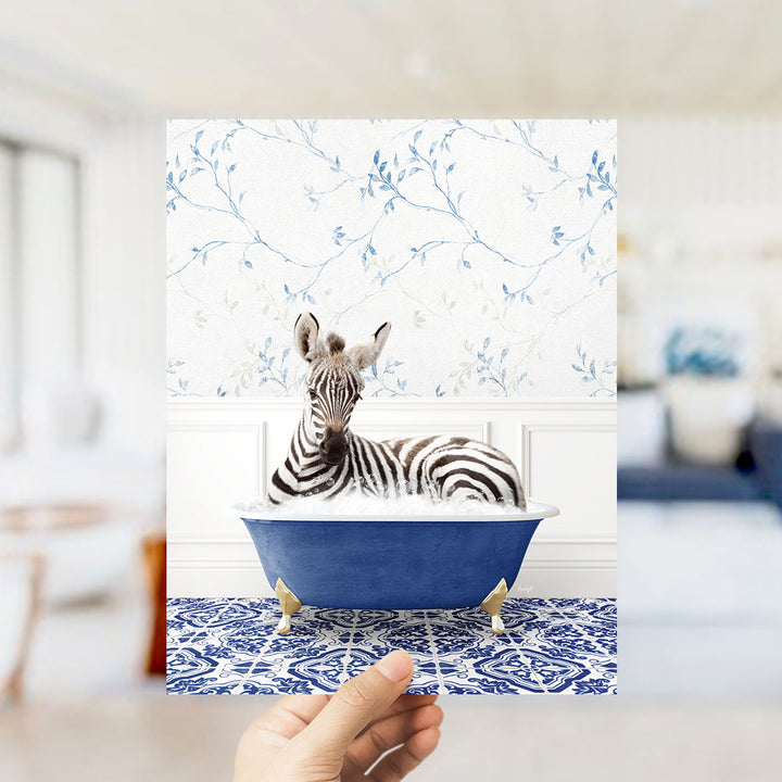 A zebra is sitting in a blue bathtub, surrounded by a patterned blue and white rug, in a room with a floral wallpaper.