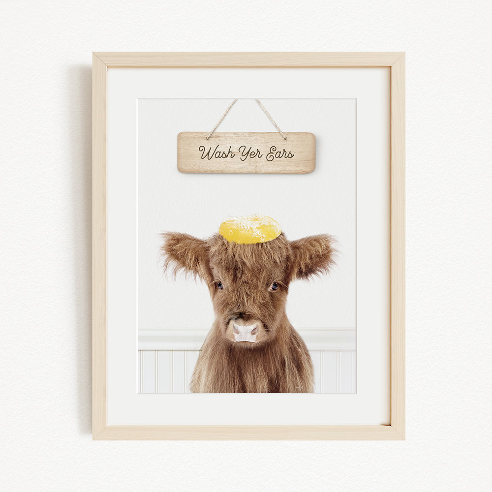A framed image of a brown cow with a yellow object on its head, accompanied by a wooden sign that reads "Wash Your Ears".
