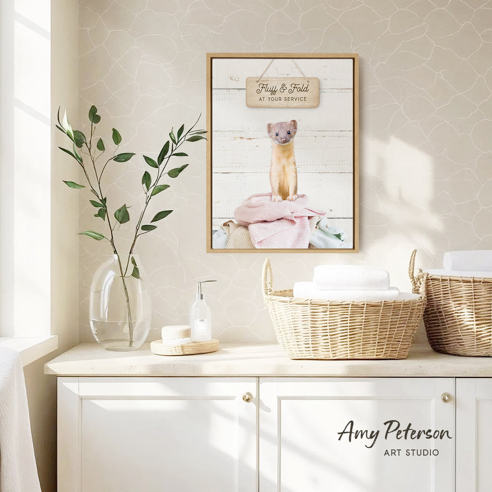 a framed picture of a small animal, likely a bird or rodent, sitting on a pink blanket in a wicker basket. The picture is hung on a wall above a white cabinet or shelf, which has a vase of green leaves and a bottle of soap on it. The cabinet also has two baskets on it, one of which is filled with towels. The overall scene appears to be a cozy and well-organized bathroom setting.