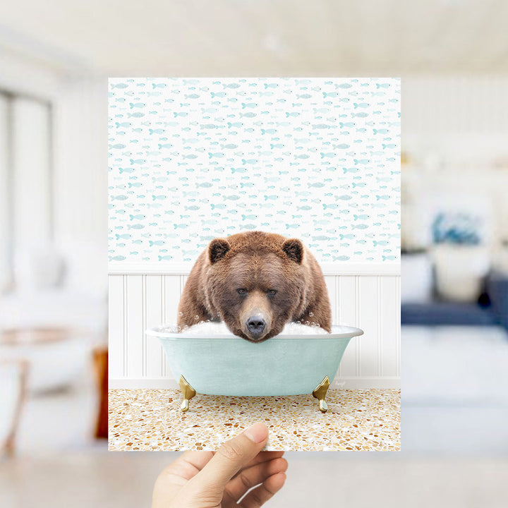 A hand holds up a framed image depicting a brown bear sitting in a bathtub filled with bubbles, surrounded by a pattern of fish.