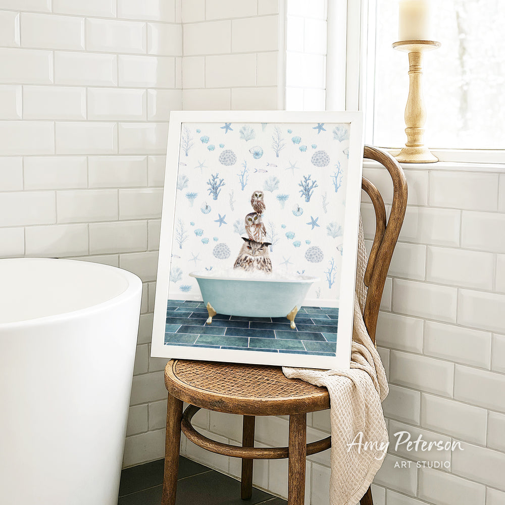 A framed image of a cat sitting in a bathtub, surrounded by blue and white seashells and starfish, is displayed on a wooden chair in a bathroom setting.