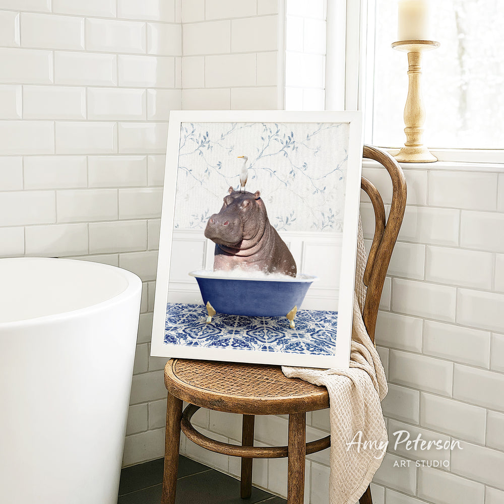 A framed image of a hippopotamus sitting in a blue bathtub, surrounded by a patterned tile floor, is displayed on a wooden chair in a bathroom setting.