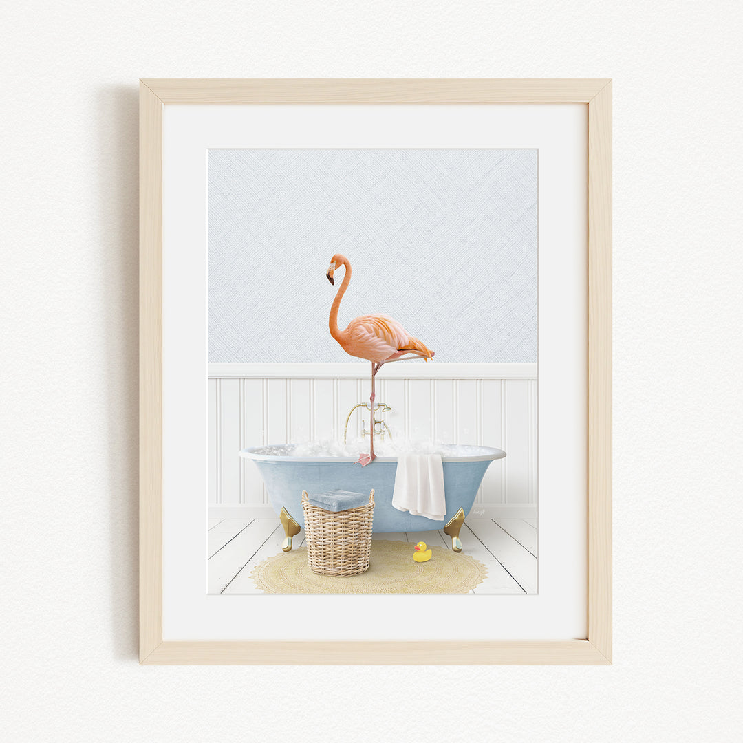 A framed image depicting a pink flamingo standing on the edge of a bathtub filled with water, surrounded by various bathroom items such as a basket, a towel, and a yellow rubber duck.