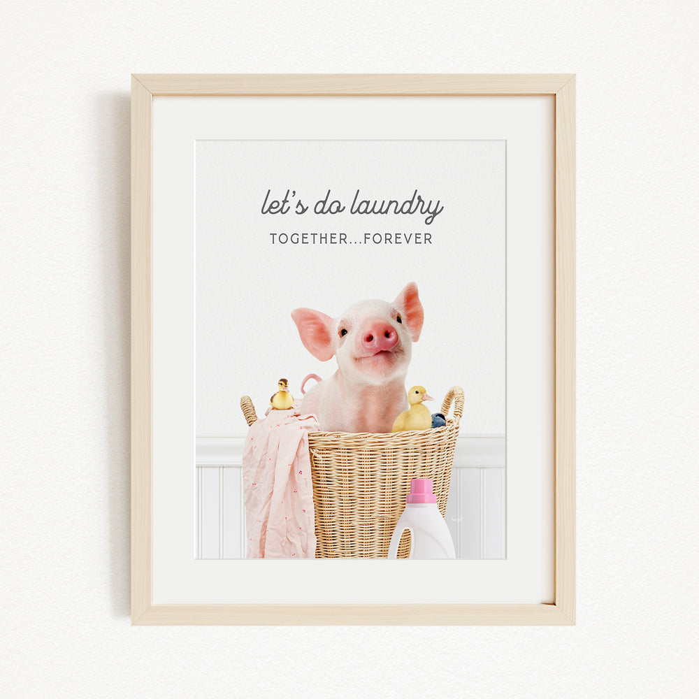 A framed image of a pig sitting in a wicker basket filled with various items, including rubber ducks and a bottle of detergent.