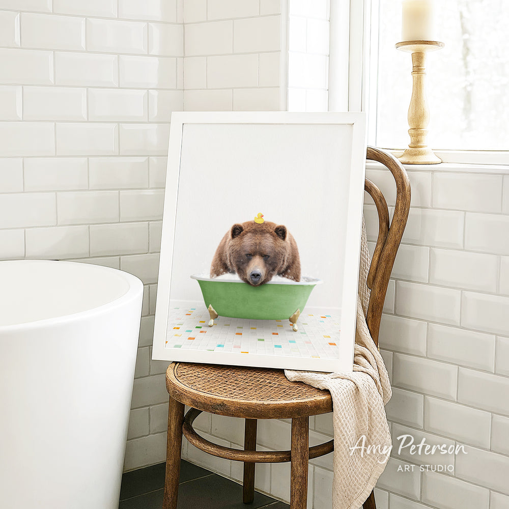 A framed image of a bear sitting in a green bathtub, with a towel draped over the back of a wooden chair in a bathroom setting.