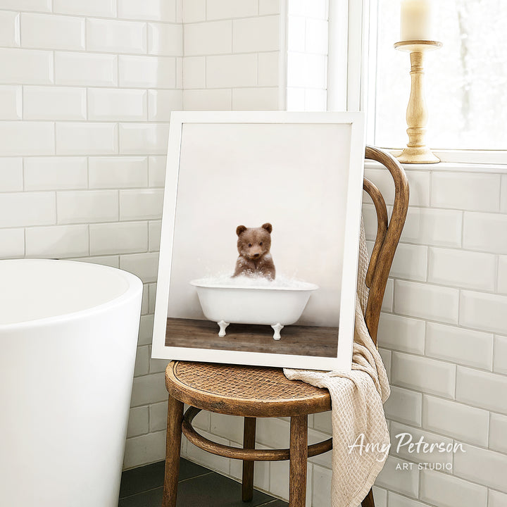 A framed image of a bear sitting in a bathtub, with a towel draped over the back of a chair in a bathroom setting.