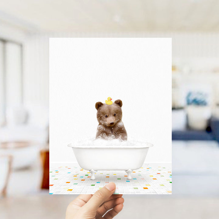 A hand holds up a framed image of a small brown bear sitting in a white bathtub filled with bubbles, with a yellow rubber duck floating on its head.