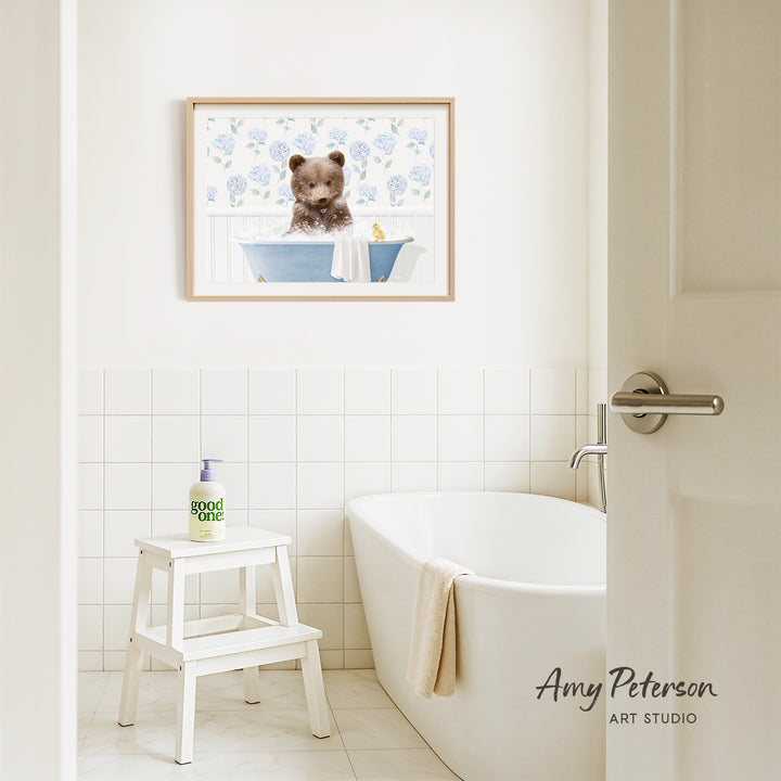 A framed image of a bear sitting in a bathtub is hanging on a white tiled wall above a white bathtub in a bathroom.