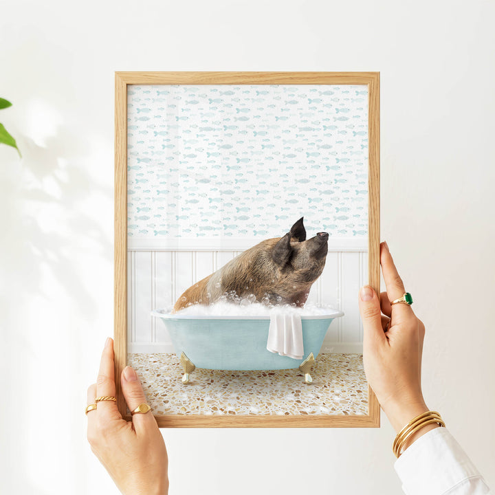A person's hands are holding a framed picture of a pig sitting in a bathtub filled with water and bubbles, with a patterned wallpaper in the background.