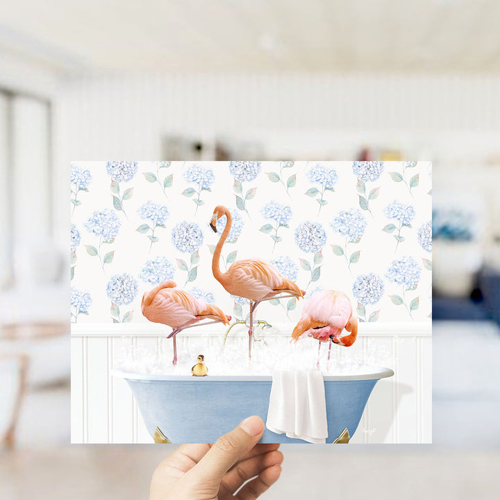 A hand holds up a framed image depicting three pink flamingos standing in a blue bathtub filled with water, surrounded by a floral pattern.