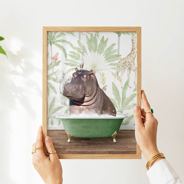 A person's hands are holding a framed picture of a hippopotamus sitting in a green bathtub, surrounded by tropical foliage.