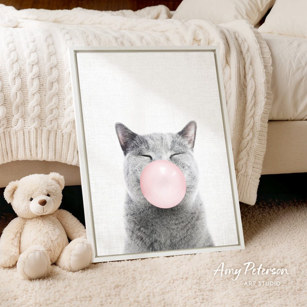 A framed image of a gray cat blowing a bubble with a pink bubble gum bubble is displayed on a white bedspread, with a teddy bear next to it.