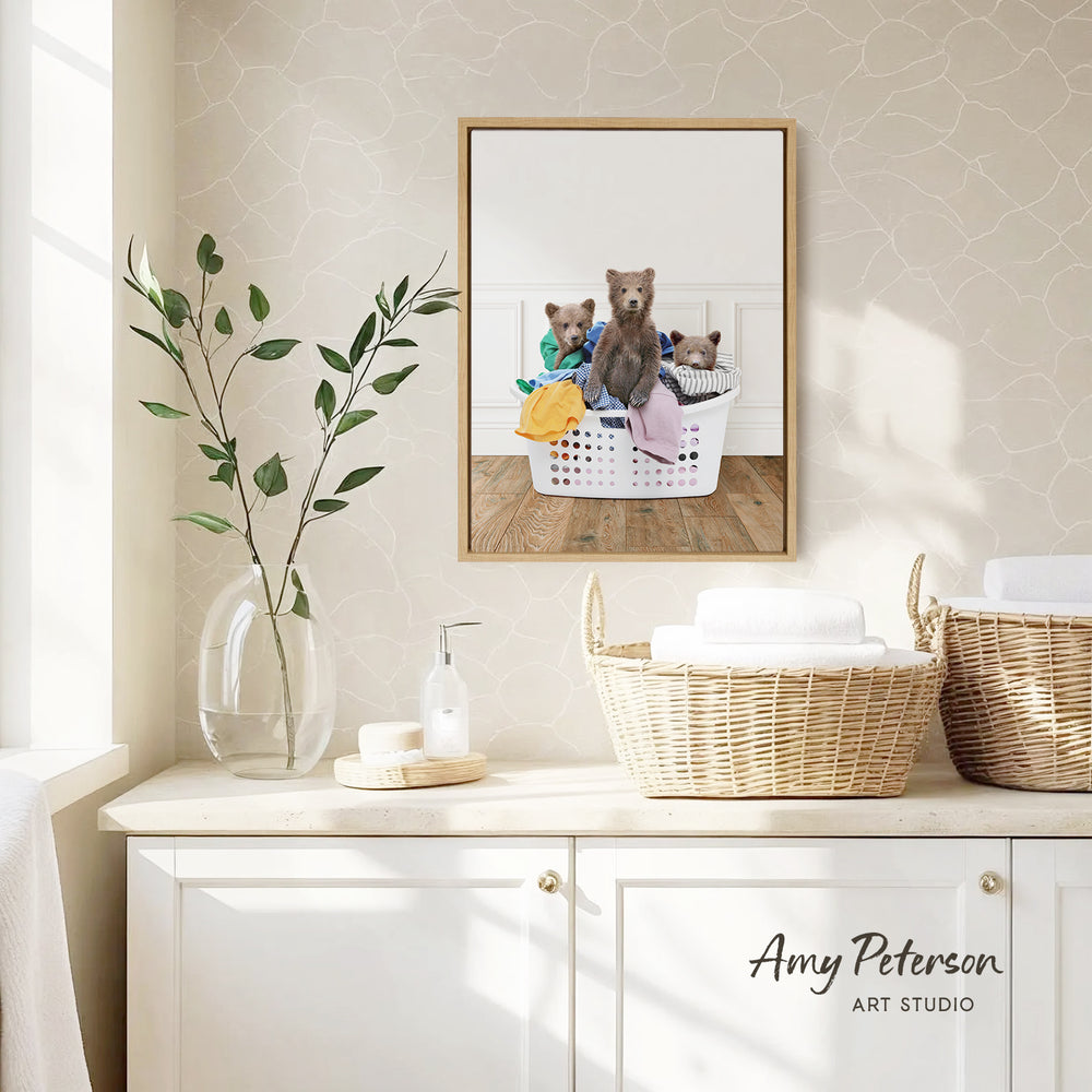 a framed photograph of three bears sitting in a laundry basket, with one bear wearing a green shirt and the other two bears wearing striped shirts. The photograph is displayed on a wall above a white cabinet with baskets and a plant.