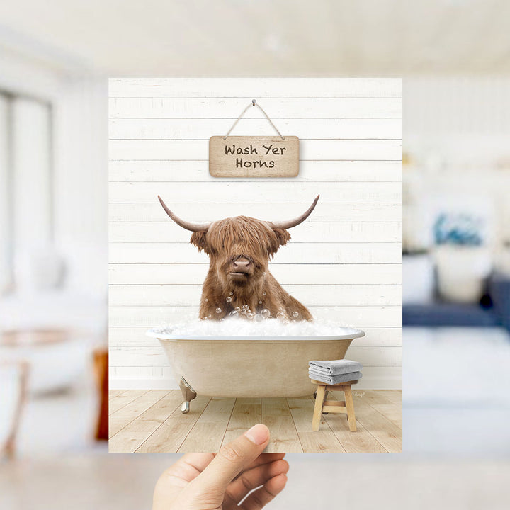 A hand holds up a framed image depicting a brown Highland cow sitting in a bathtub, with a sign above that reads "Wash Yer Horns".