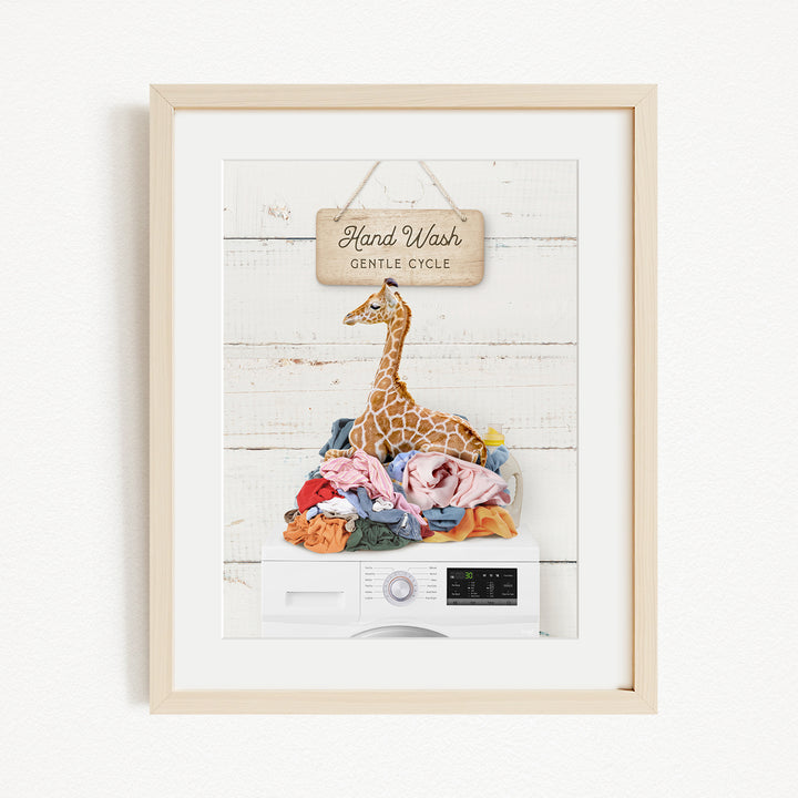 A framed image depicting a giraffe sitting atop a pile of laundry, with a sign above that reads "Hand Wash Gentle Cycle".