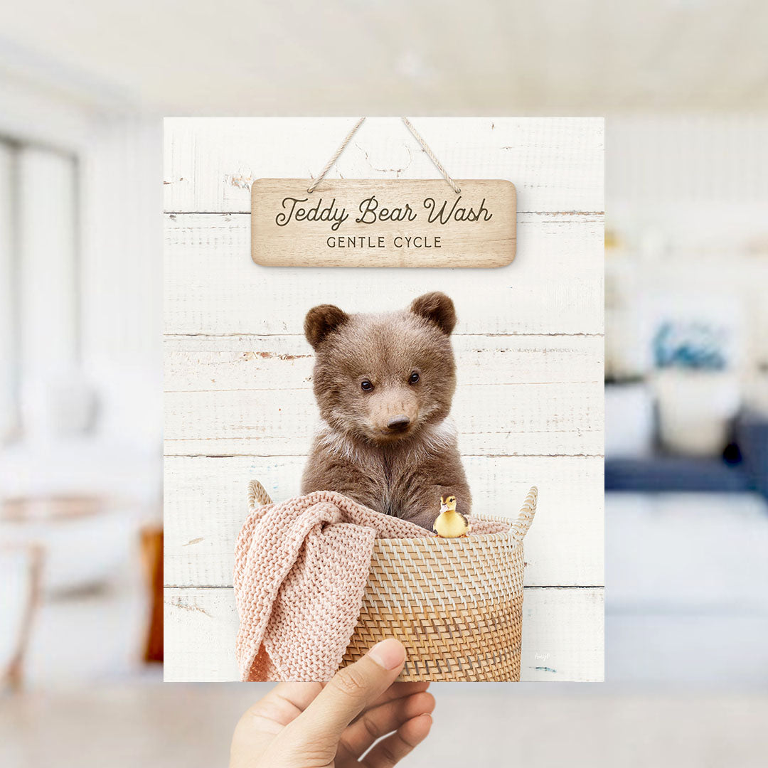 A hand is holding up a framed image of a teddy bear sitting in a woven basket, with a sign above it that reads "Teddy Bear Wash Gentle Cycle".