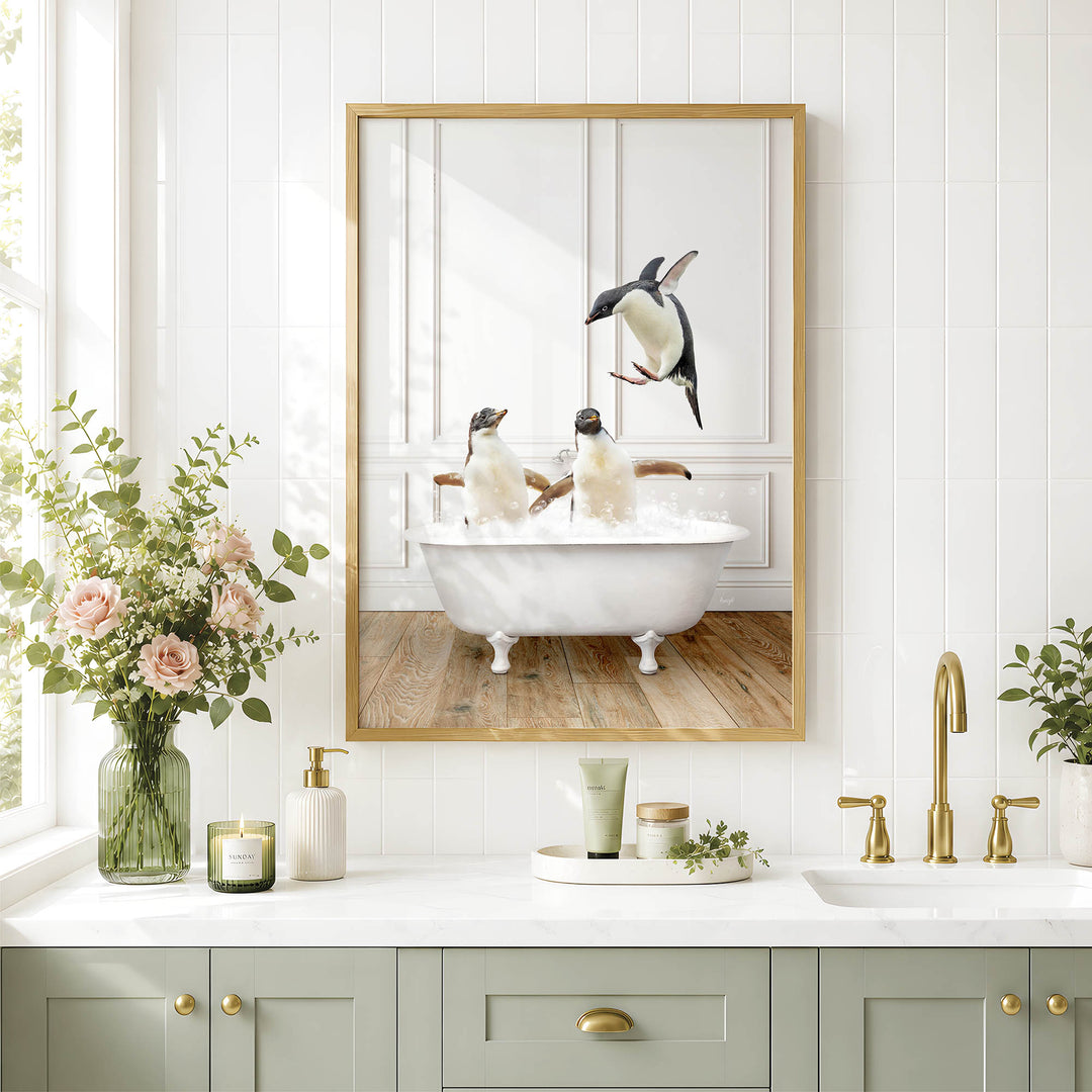 A framed image of two penguins sitting in a bathtub, surrounded by a vase of flowers, a candle, and a small plant on a white countertop in a bathroom setting.