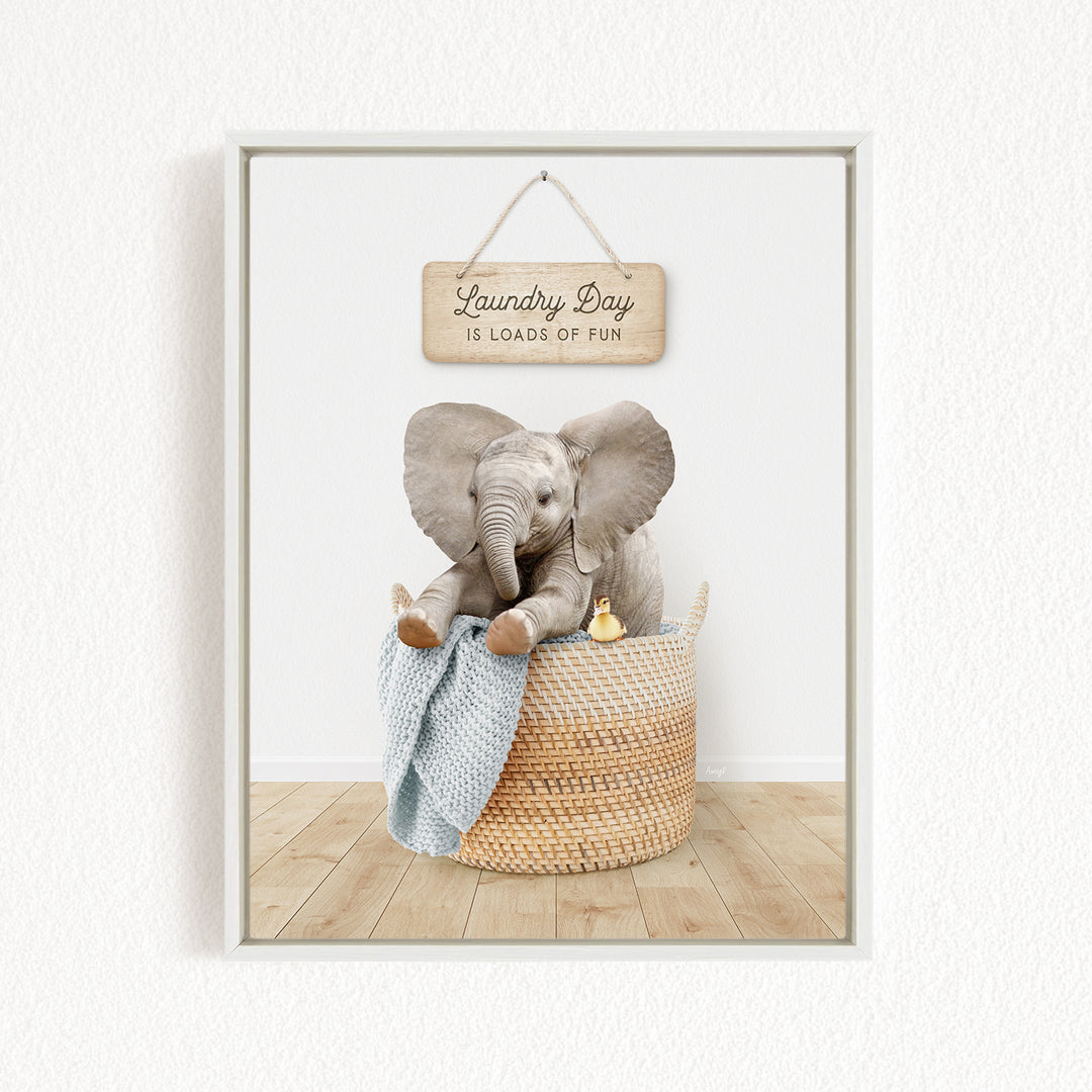 A framed image of a baby elephant sitting in a woven basket, with a sign above that says "Laundry Day is loads of fun".