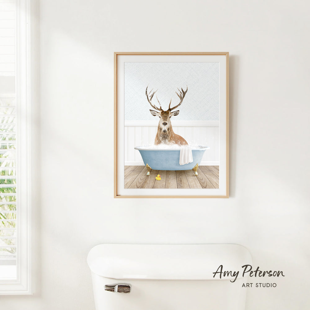 A framed print of a deer sitting in a bathtub is hanging on a white wall.