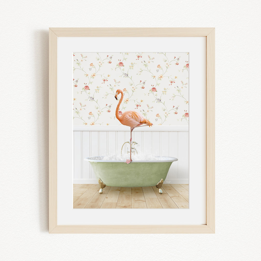 A framed image depicting a pink flamingo standing on the edge of a green bathtub filled with water, set against a floral wallpaper background.