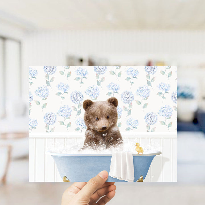 A hand holding a framed image of a brown bear sitting in a blue bathtub, surrounded by blue hydrangea flowers.