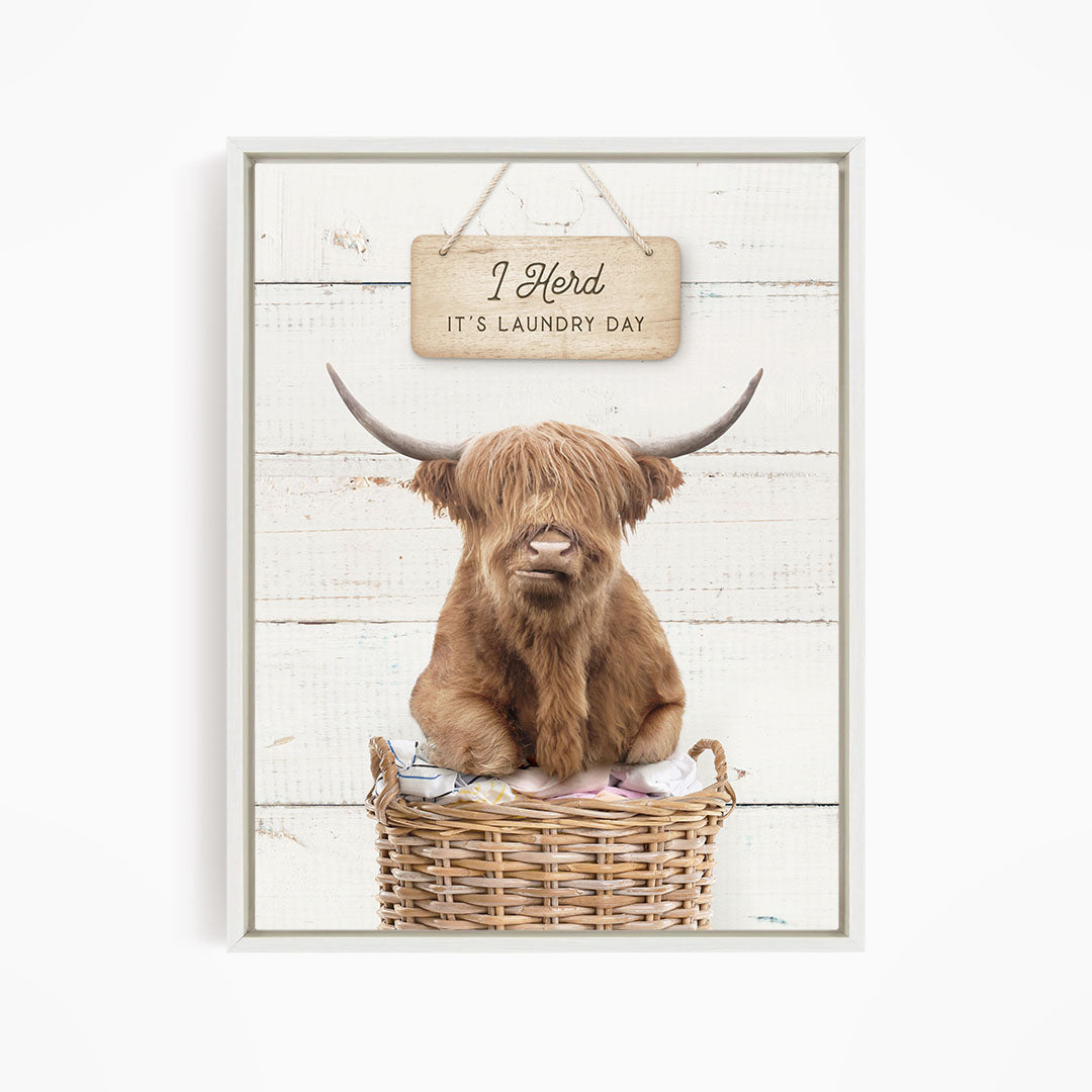 A brown Highland cow with shaggy hair sits in a wicker basket, with a wooden sign above it that says "It's laundry day".
