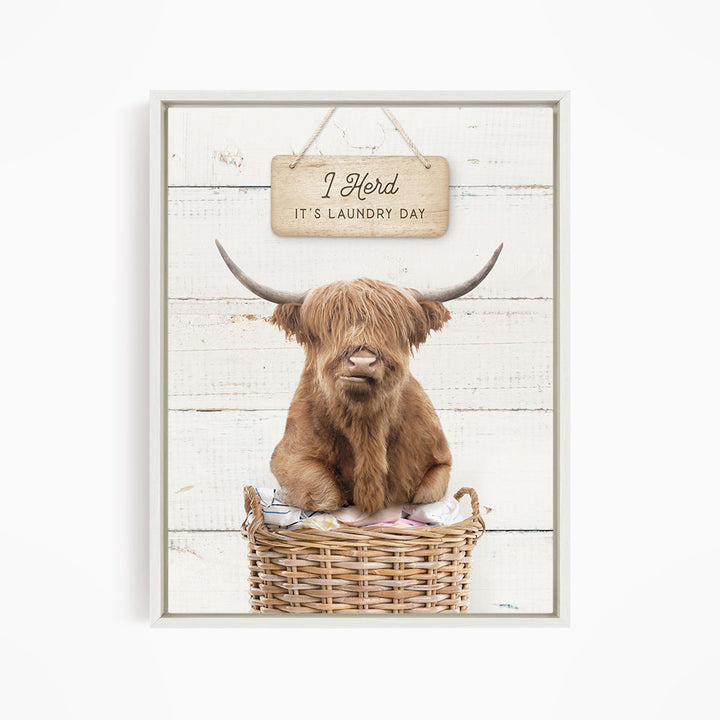 A brown Highland cow with shaggy hair sits in a wicker basket, with a wooden sign above it that says "It's laundry day".
