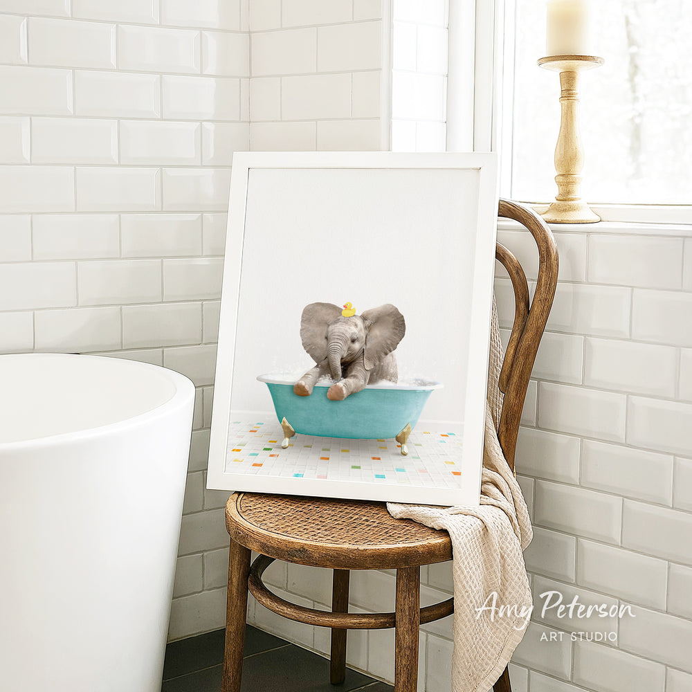A framed image of a baby elephant sitting in a blue bathtub, surrounded by colorful tiles, is displayed on a wooden chair in a bathroom setting.