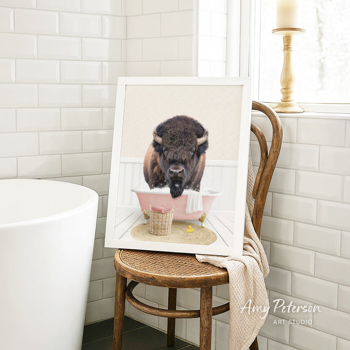 A framed image of a buffalo sitting in a pink bathtub, with a towel and basket nearby, is displayed on a wooden chair in a bathroom setting.