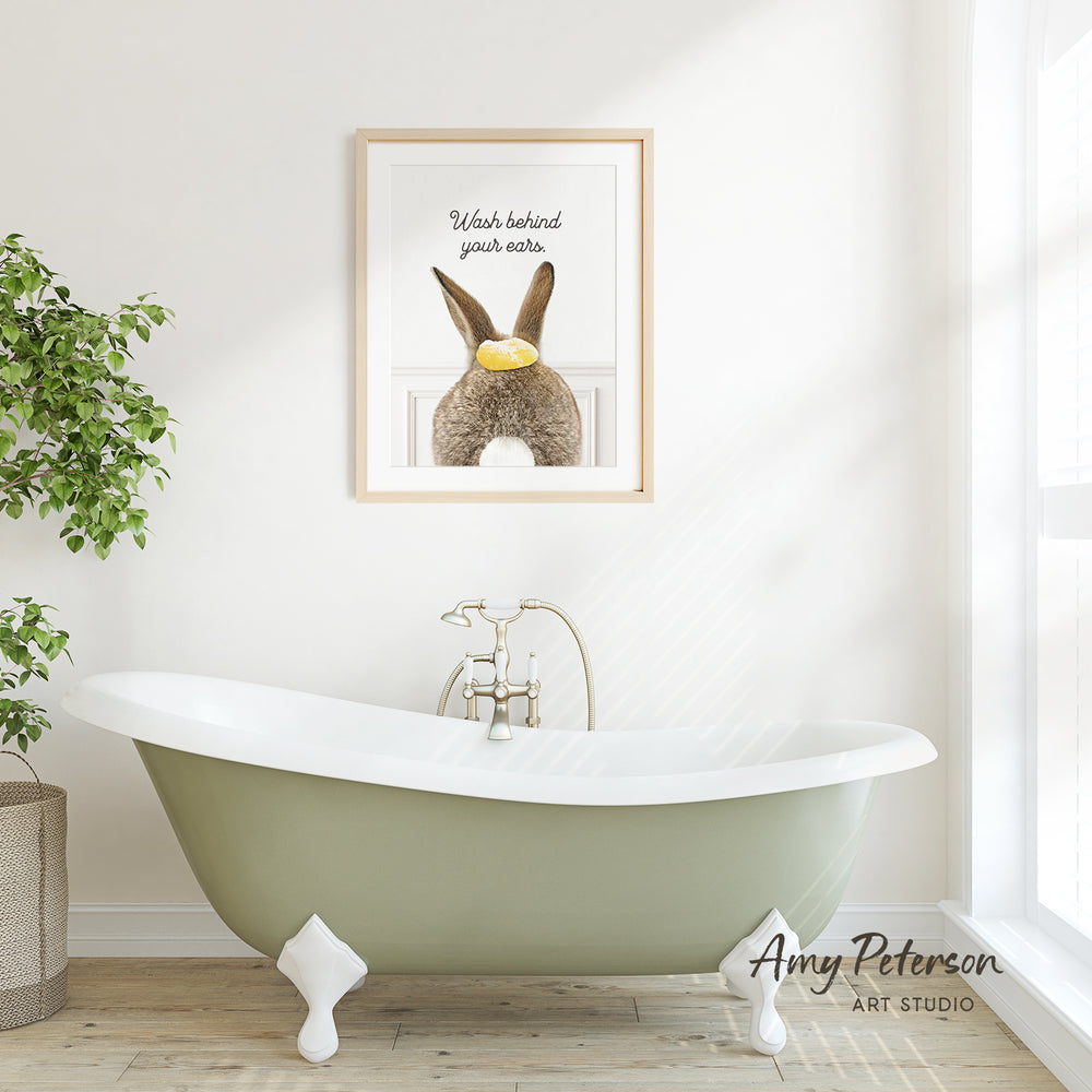 A framed print of a rabbit with a yellow object in its mouth hangs on a white wall above a green claw-foot bathtub in a bright, airy bathroom.