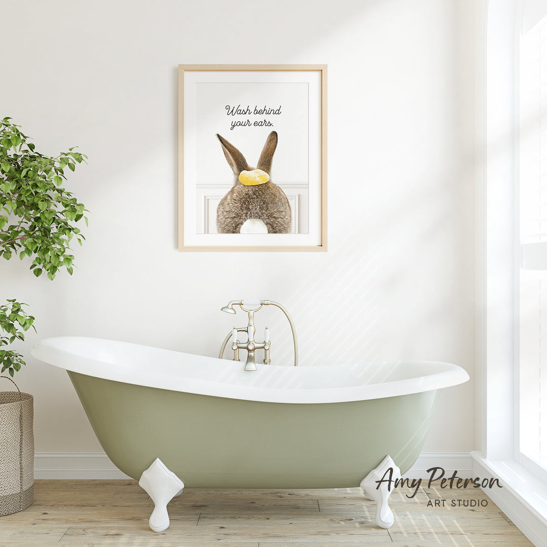 A framed print of a rabbit with a yellow object in its mouth hangs on a white wall above a green claw-foot bathtub in a bright, airy bathroom.