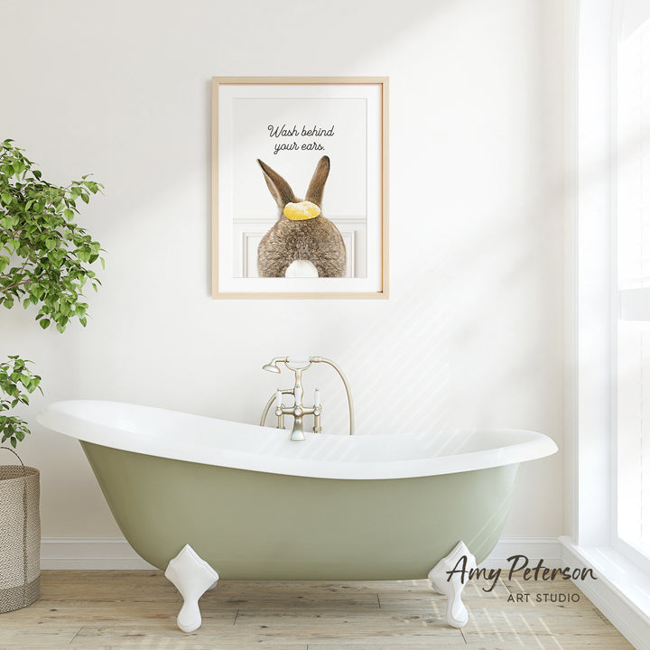 A framed print of a rabbit with a yellow object in its mouth hangs on a white wall above a green claw-foot bathtub in a bright, airy bathroom.