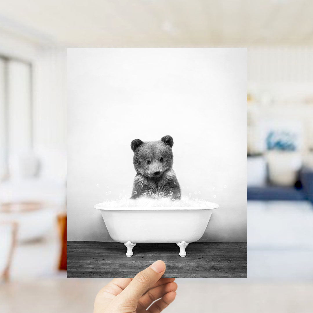 A hand holds up a black and white photograph of a baby bear sitting in a bathtub filled with water, creating a whimsical and playful scene.
