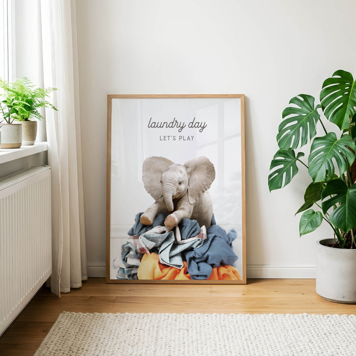 A framed poster with an elephant sitting on a pile of clothes, accompanied by the text "laundry day let's play".