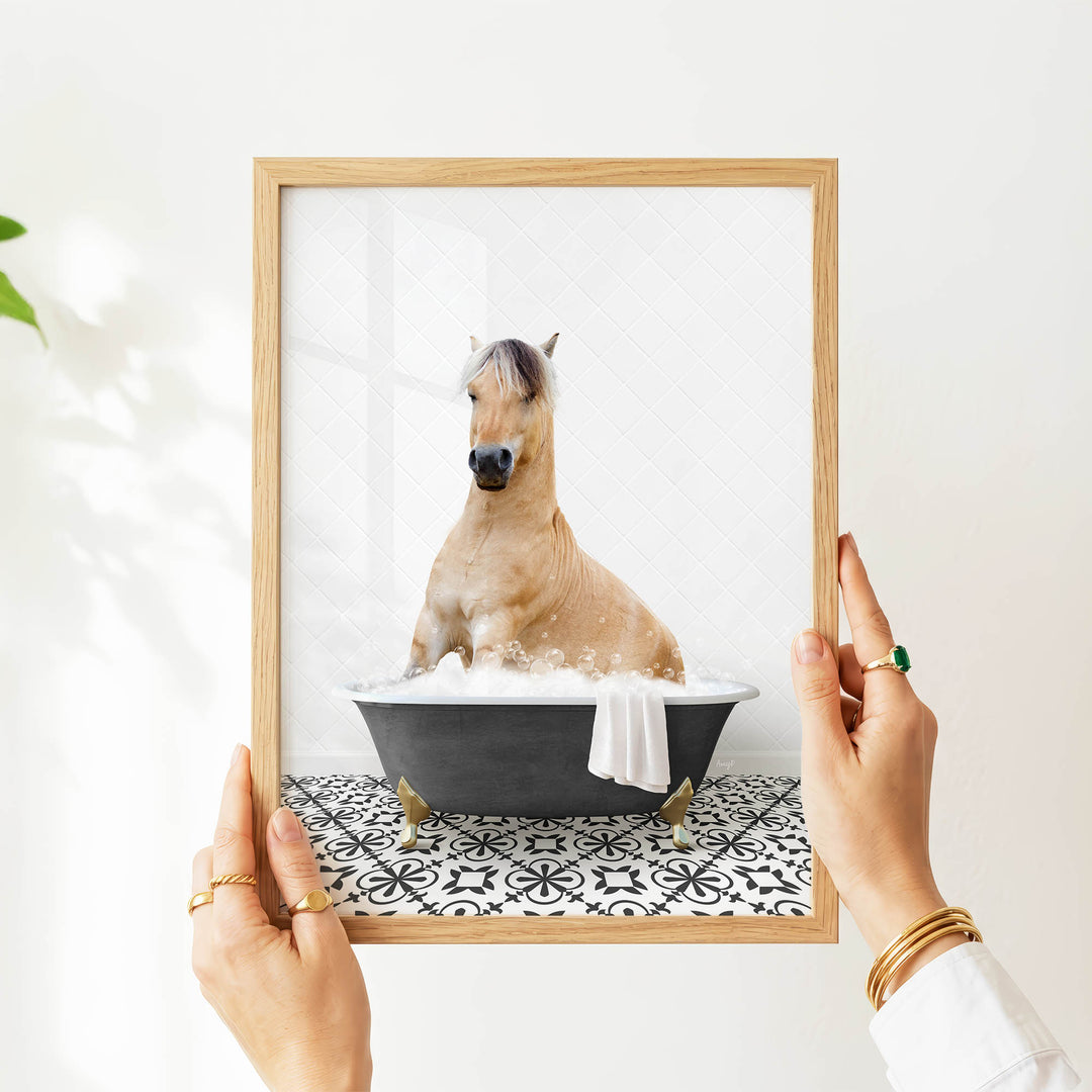 A person's hands are holding a picture frame containing an image of a horse sitting in a bathtub filled with water and bubbles.